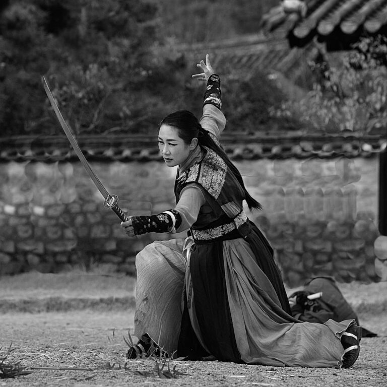 A woman in Korean traditional dress kneeling with a raised sword in the courtyard of a hanok