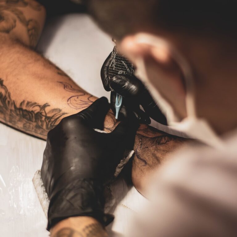 A tattoo artist in black gloves working on a client's forearm, seen over the shoulder