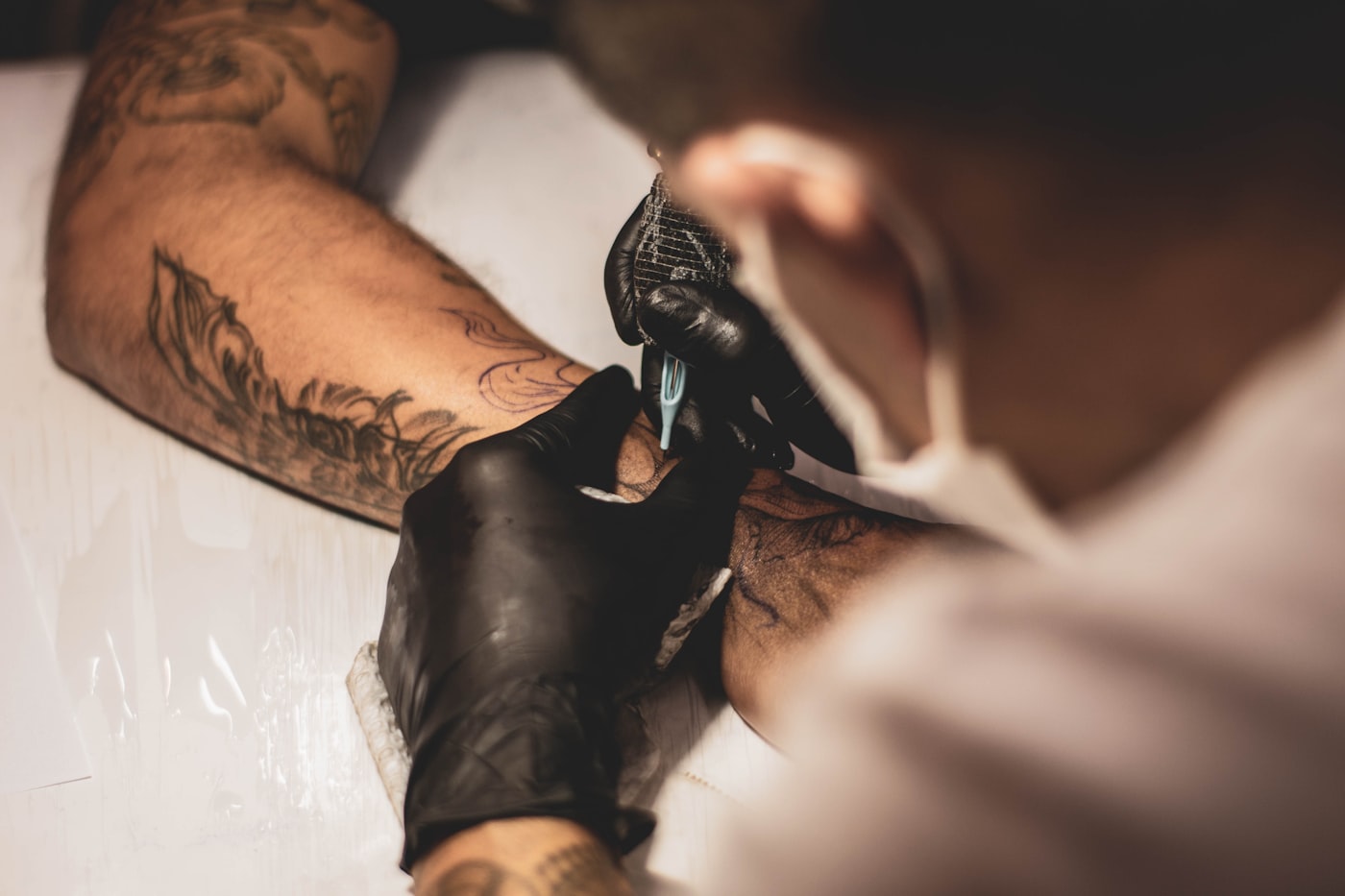 A tattoo artist in black gloves working on a client's forearm, seen over the shoulder